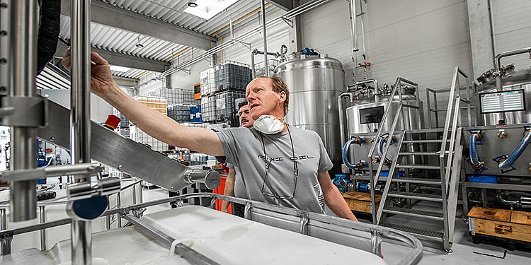 Die Szene zeigt eine industrielle Umgebung mit großen Tanks und einem Mann, der eine Abfüllmaschine bedient.