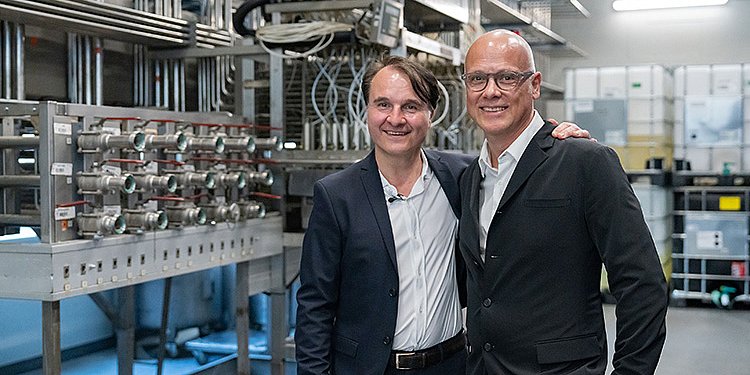 Zwei Männer in Anzügen stehen lächelnd in einer modernen Produktionsumgebung mit Maschinen im Hintergrund.