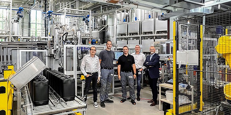 Die Gruppe von fünf Männern steht in einer modernen Fabrik, umgeben von Abfüllanlagen und technischen Geräten.