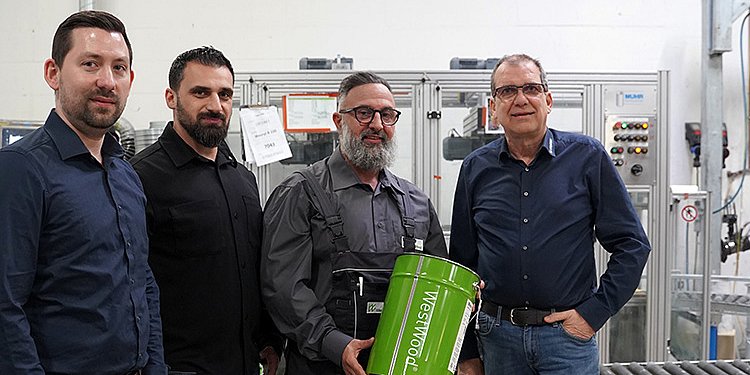 Mitarbeiter von Fricke und Westwood stehen vor einer Abfüllanlage und eine Person hält einen Eimer mit Westwood Schriftzug.