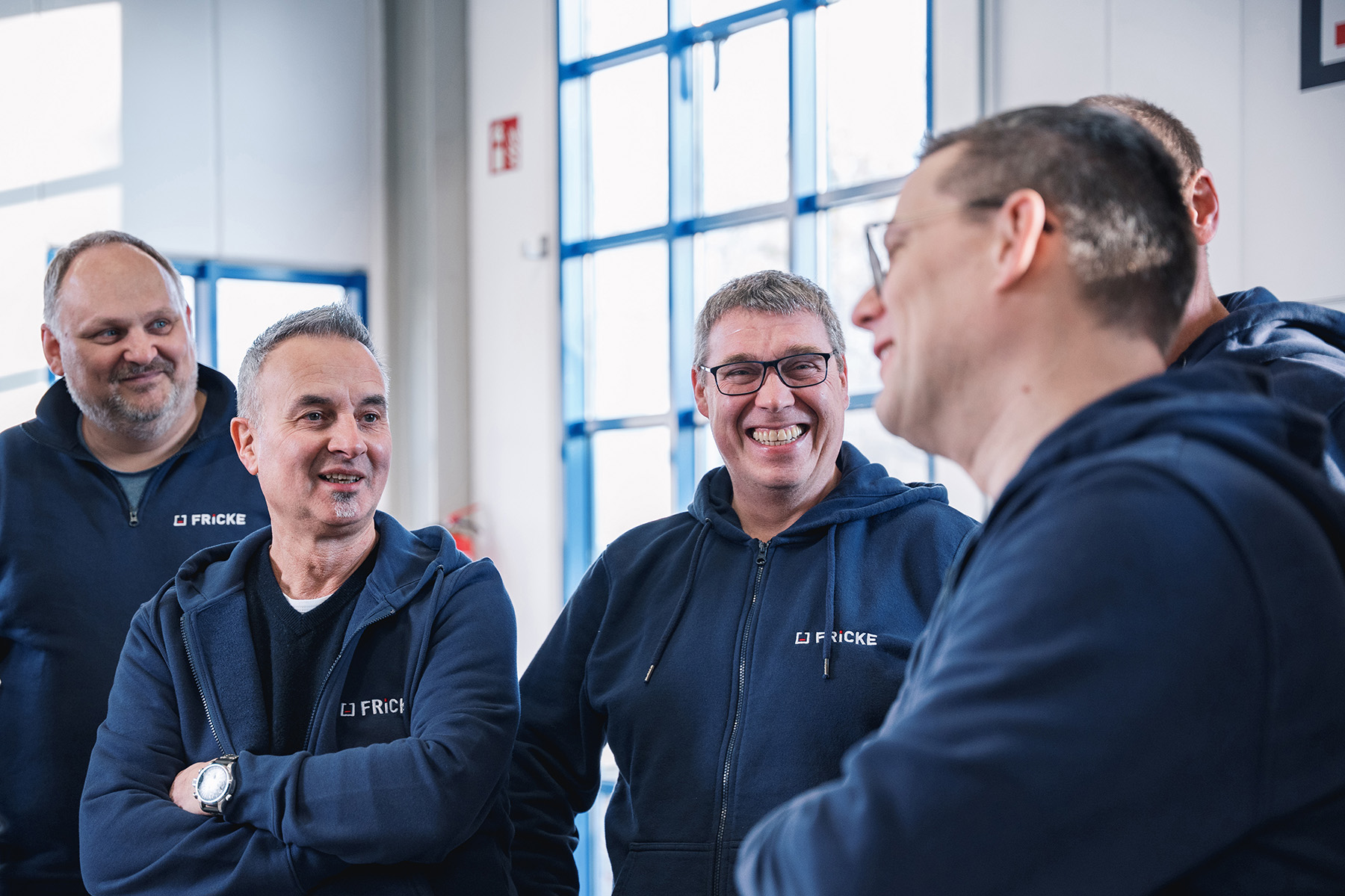 Several men wearing FRICKE clothing are standing in a bright room and engaged in lively discussion.