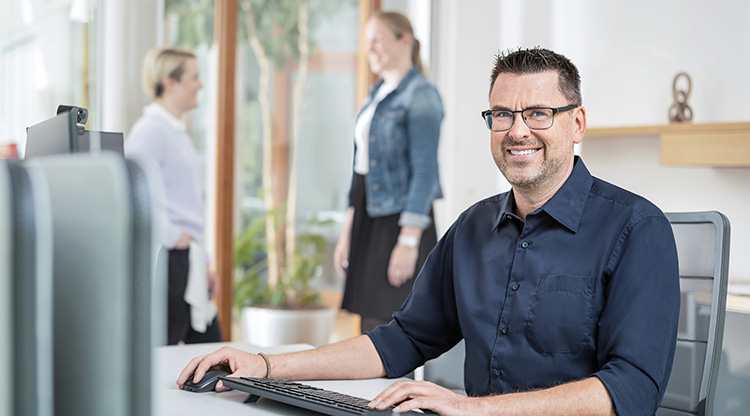 A smiling man sits at a desk working on a computer while two women talk to each other in the background.