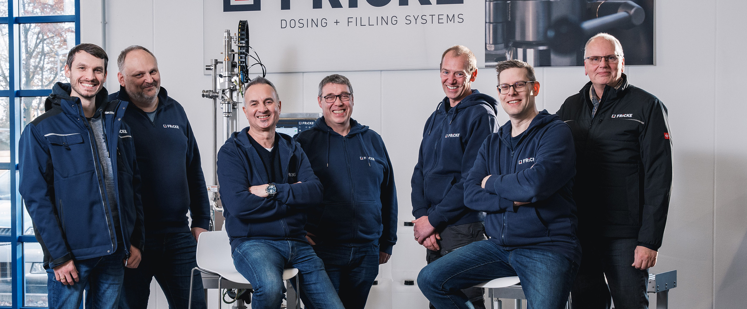The cheerful faces of FRICKE employees radiate team spirit as they stand together in a modern working environment.