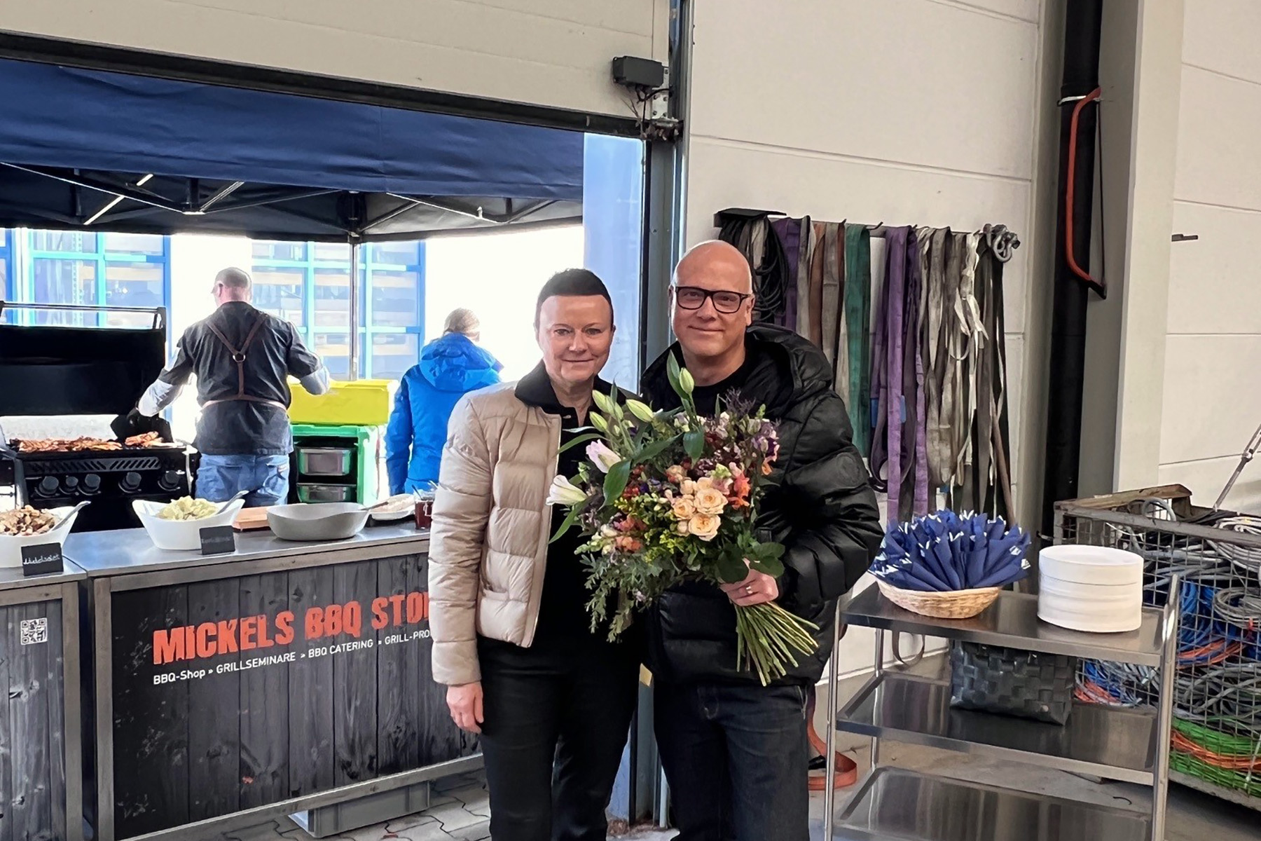 In the background, someone is grilling meat, while the couple in the foreground smiles at the camera with a bouquet of flowers.