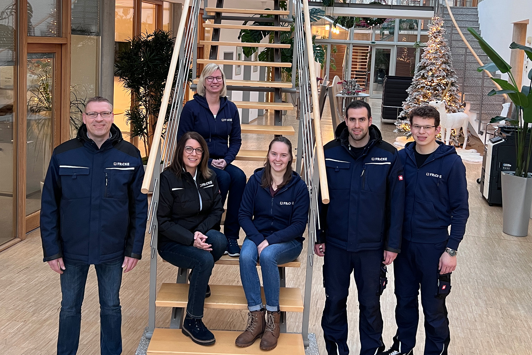 A group of employees stands on a staircase, surrounded by plants and festive Christmas decorations in the bright FRICKE foyer.