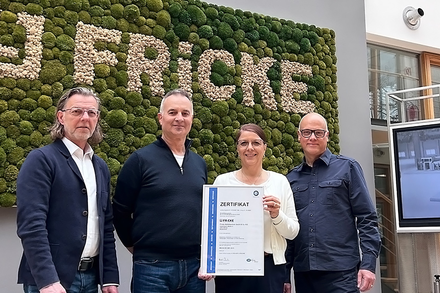 The group proudly displays a certificate while posing in front of a vibrant moss wall that spells out the company name FRICKE.