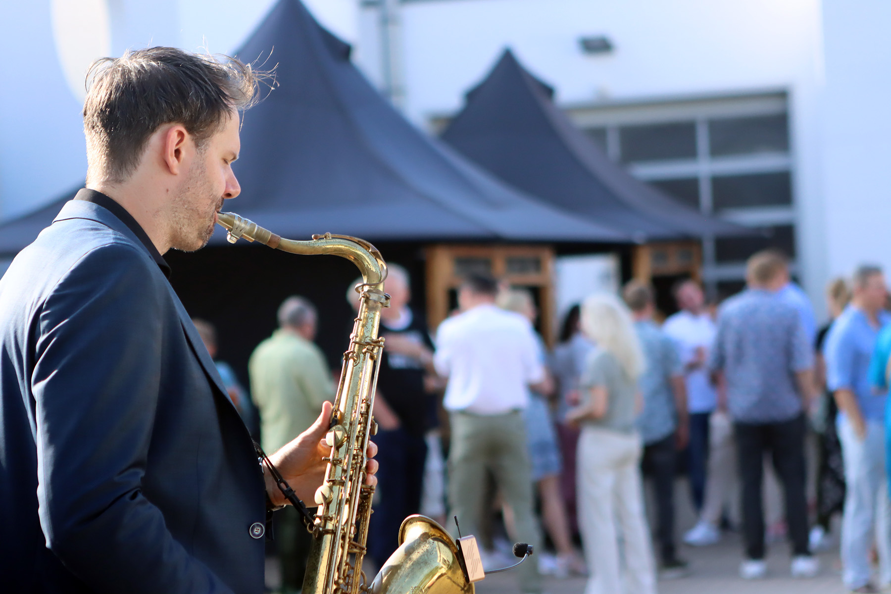A saxophonist plays passionately in a festive atmosphere while a group of people celebrate in the background.