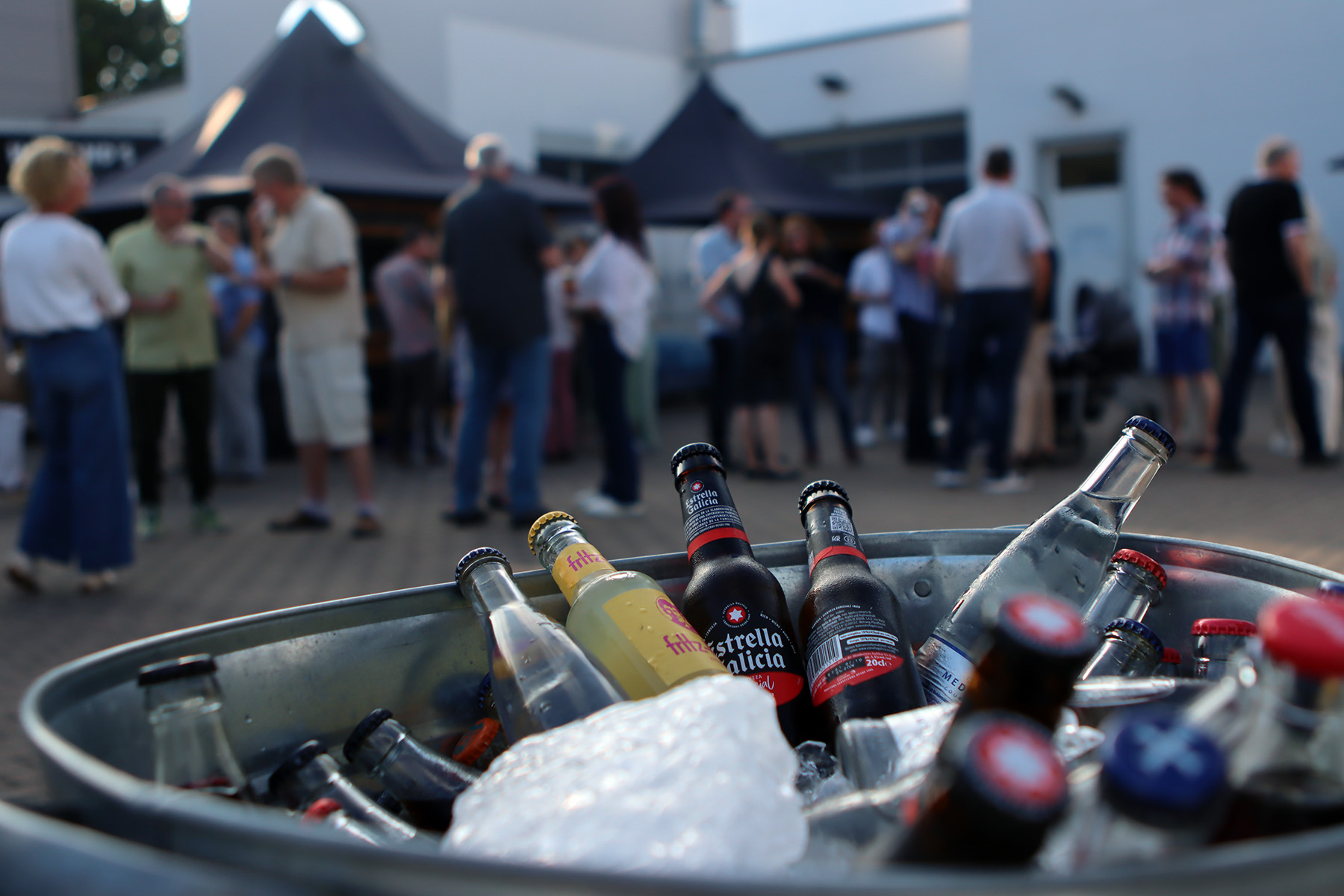 A variety of drinks in a cooler, while cheerful guests attend a party and chat in the background.