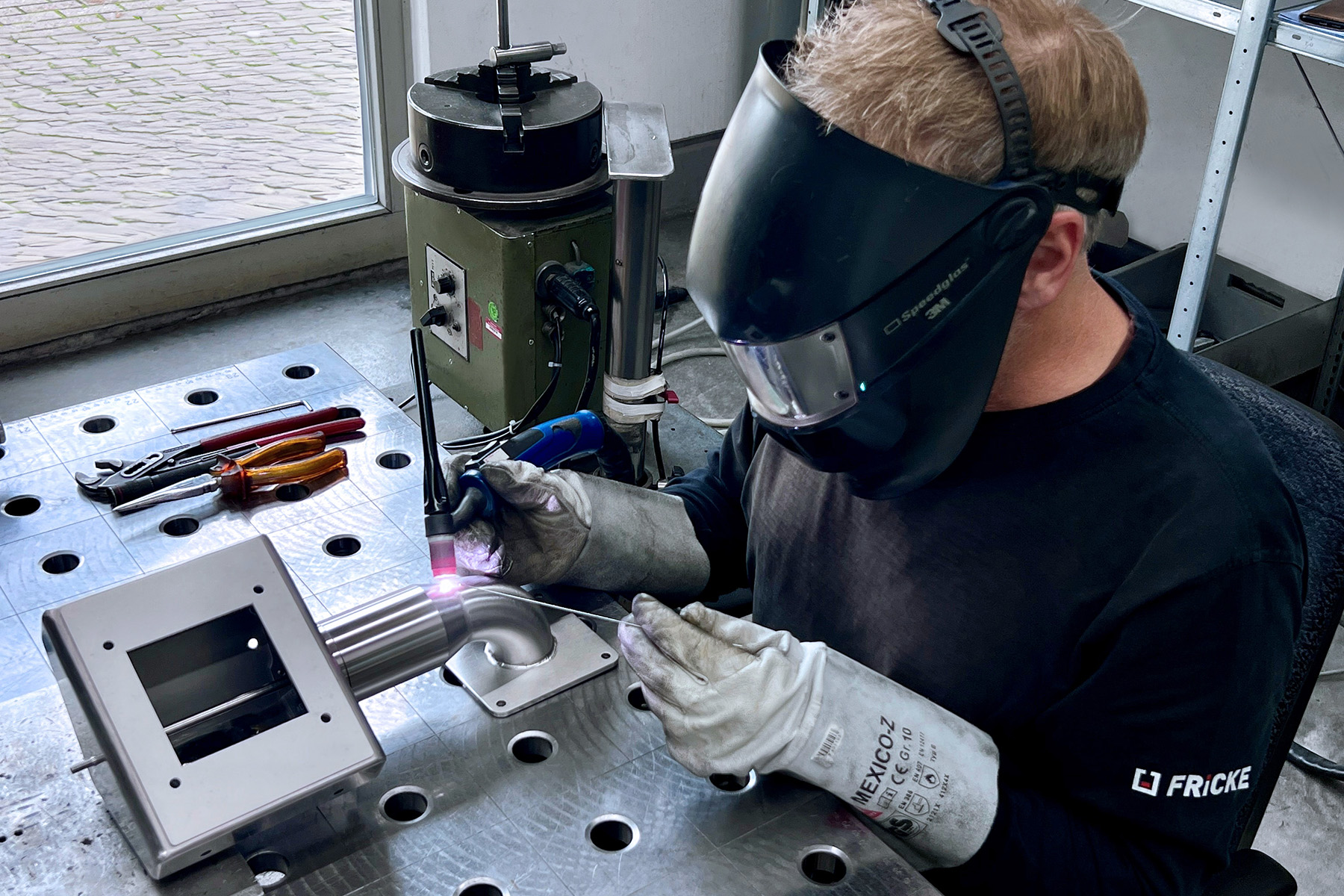 A welder works intently on a piece of metal, holding a welding torch in his hand and wearing protective clothing.
