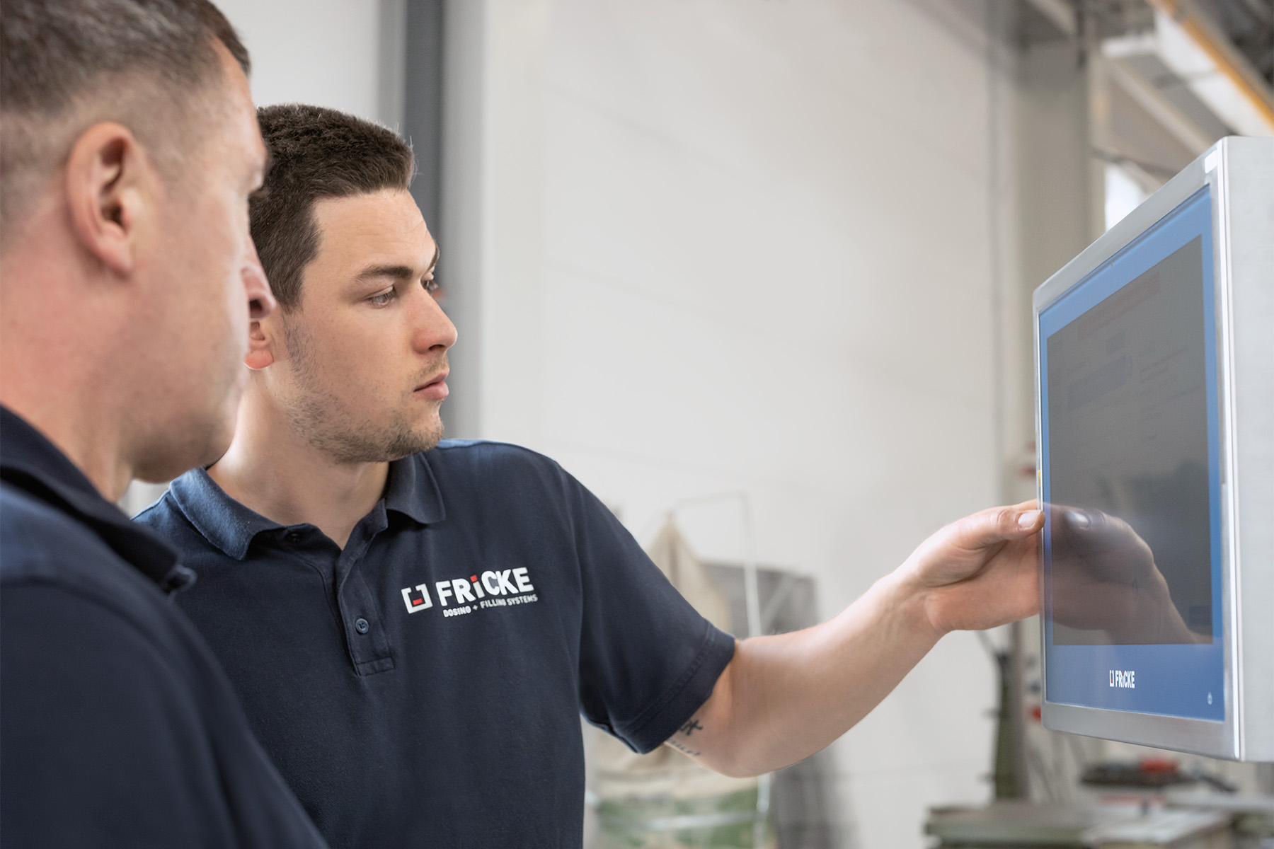 A young man points to a screen while a colleague listens attentively; both are wearing polo shirts with the ‘FRICKE’ logo.