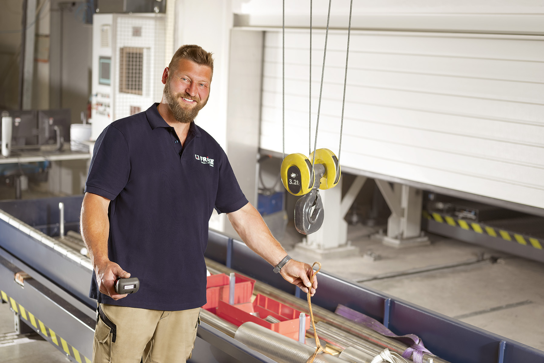 In a workshop, a man smiles as he holds a remote control in his hand and points to a lifting device.