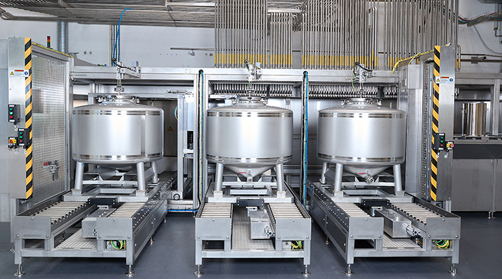 Drei große, silberne Behälter stehen auf Förderbändern in einer modernen Produktionsanlage, umgeben von industrieller Ausrüstung.