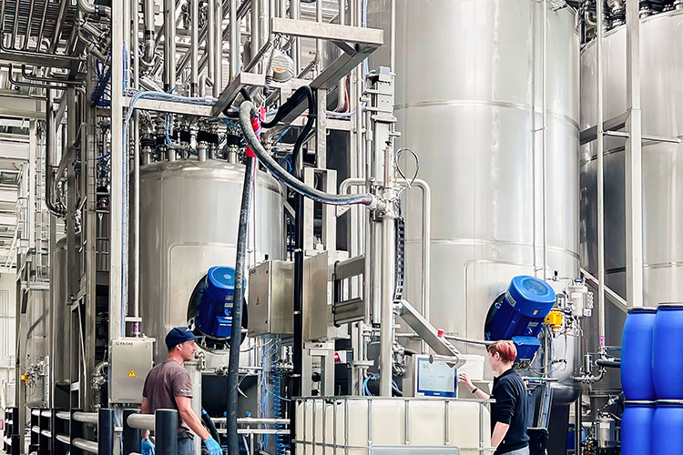 Zwei Mitarbeiter arbeiten an einer modernen Abfüllanlage, umgeben von großen silbernen Tanks und blauen Fässern.