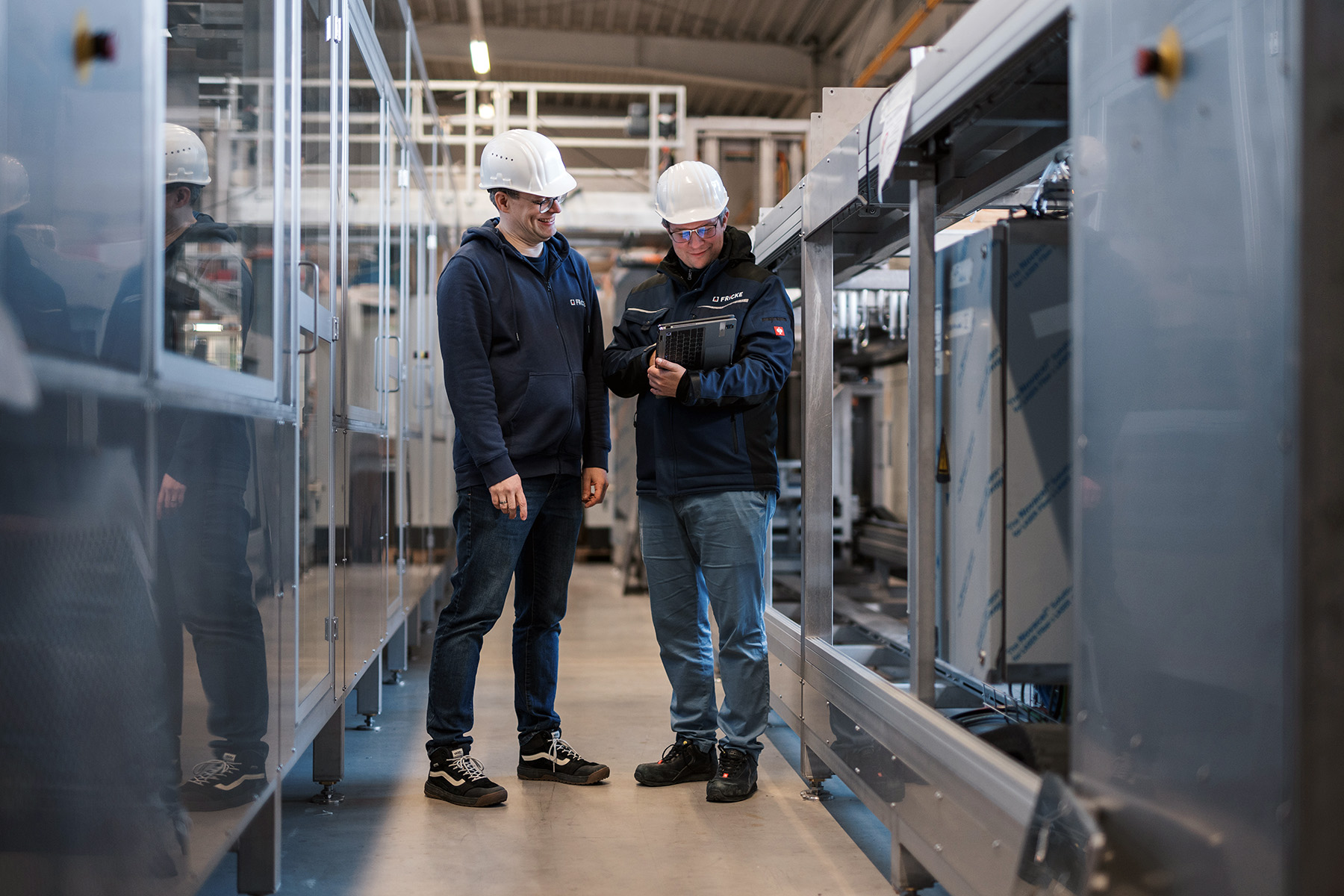 Zwei Männer in Schutzhelmen stehen in einer modernen Produktionshalle und betrachten ein Tablet mit technischen Informationen.