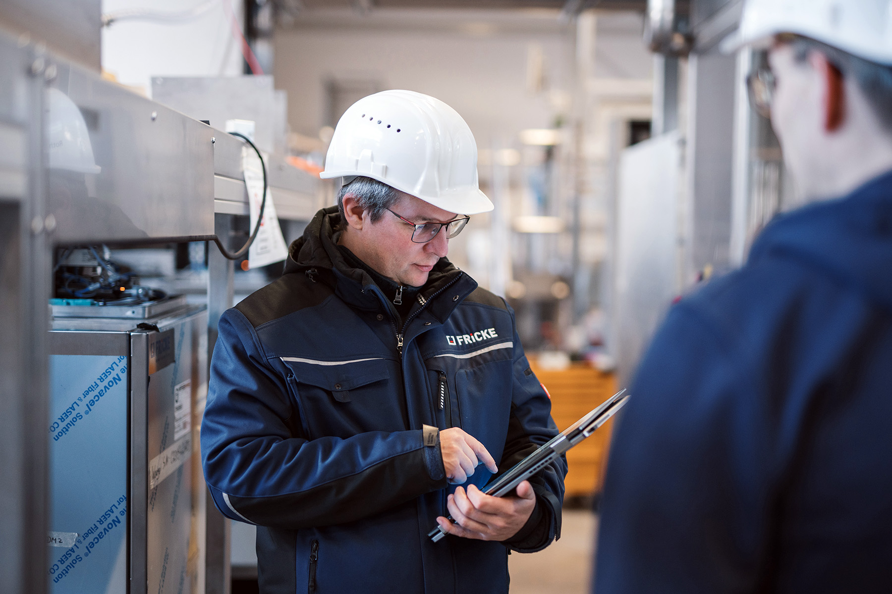 In einer industriellen Umgebung analysiert ein Mann mit Brille und Helm Daten auf einem Tablet, während er konzentriert wirkt.