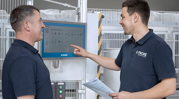 Two men stand in front of a screen and discuss the operation of the dosing system in a modern production environment.