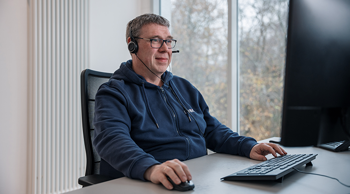 A man wearing glasses and a headset sits at a desk, concentrating on his work at a computer.