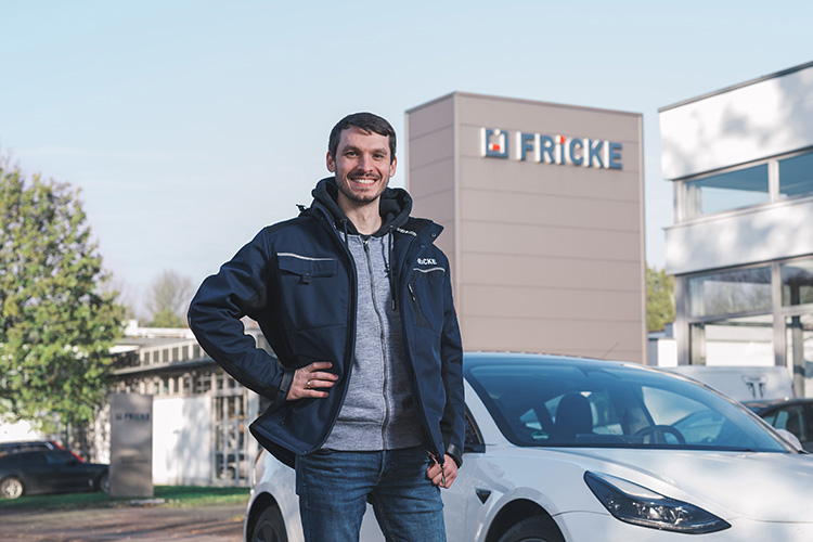 Ein lächelnder Mann in einem blauen Arbeitsjacke steht vor einem modernen Gebäude mit dem Logo 'FRICKE' und einem weißen Auto.
