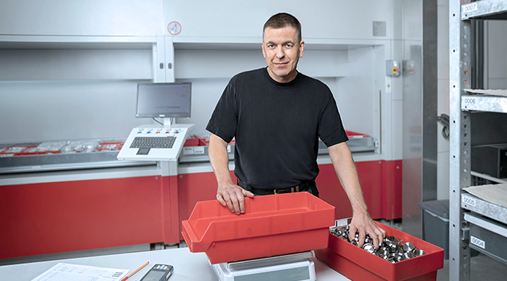 In a storage room, a man is sorting spare parts, surrounded by shelves and a computer.